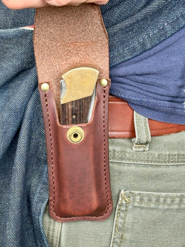 Brown leather knife sheath with brass fasteners worn on a person's waist.