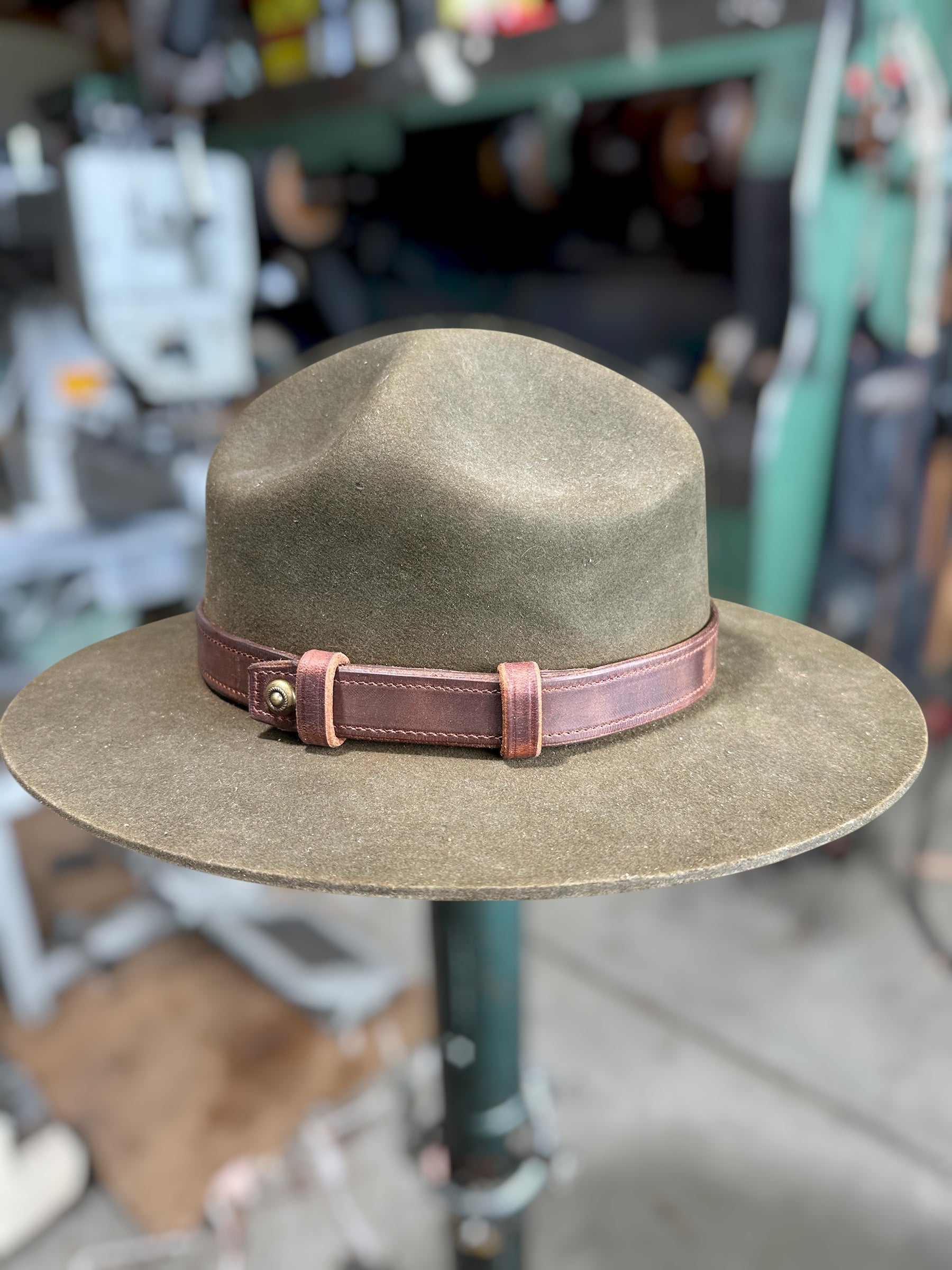 A wide-brim fedora hat in olive brown felt with a dusty rose leather band and brass buckle, displayed on a stand against a blurred workshop background.