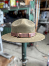 A wide-brim fedora hat in a muted taupe-brown color, featuring a contrasting dusty rose pink leather band with a brass snap closure, displayed on a stand in a workshop setting.