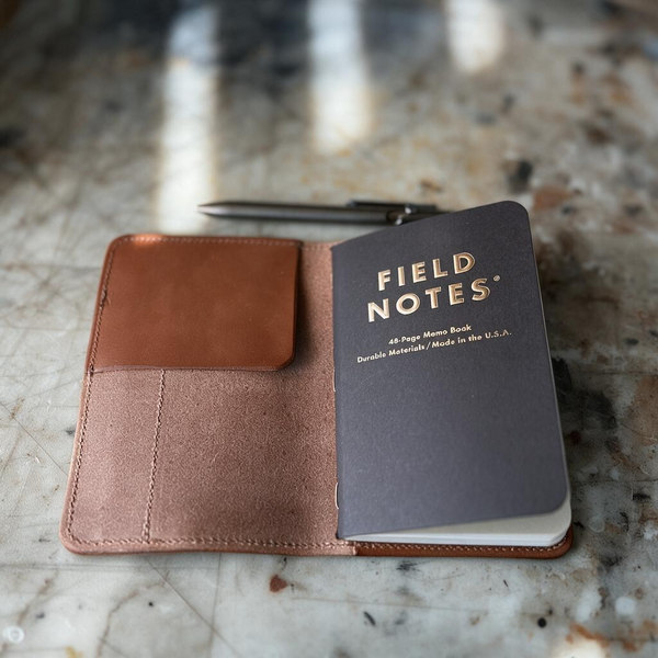 An open brown leather notebook cover on a marble surface, holding a dark gray Field Notes 44-page home notebook and a black pen.