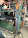 A close-up of black leather belt being held by a hand, showing the buckle, strap, and a stamped logo on the keeper loop. The background is a blurred workshop environment.