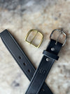 A black leather belt laid on a light stone surface, shown with a gold-toned square buckle and a silver-toned rectangular buckle.