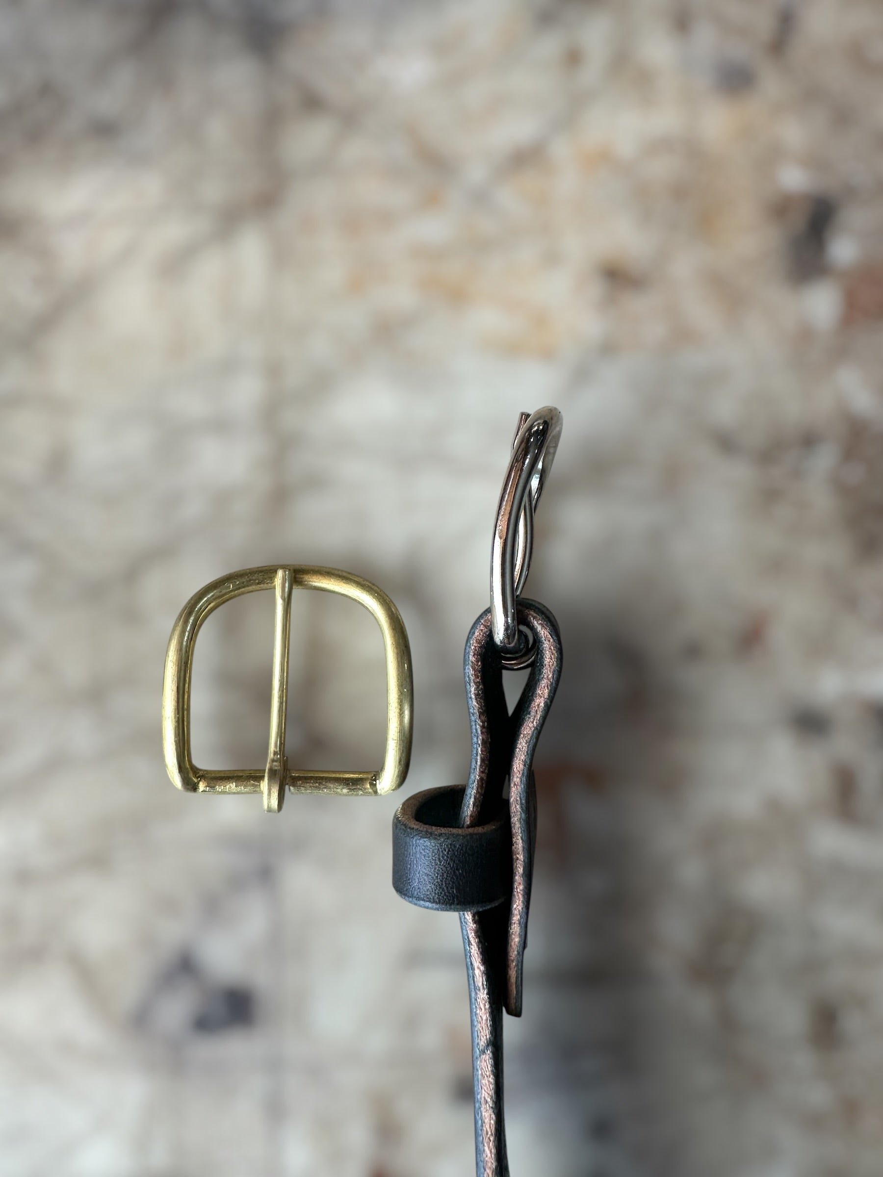 A close-up of a brass buckle and a dark brown leather strap with a silver metal loop, set against a blurred beige stone background.