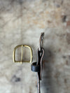 A close-up of a brass buckle and a dark brown leather strap with a silver metal loop, set against a blurred beige stone background.