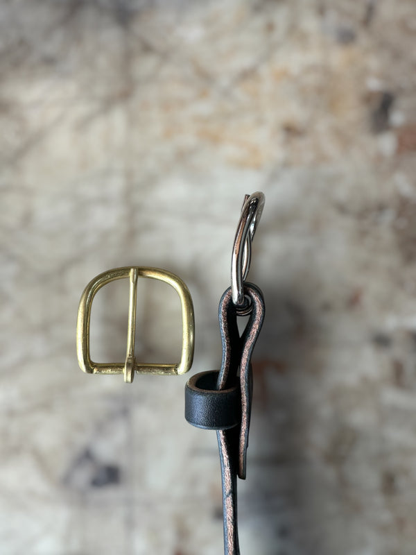 A close-up of a brass buckle and a dark brown leather strap with a silver metal loop, set against a blurred beige stone background.