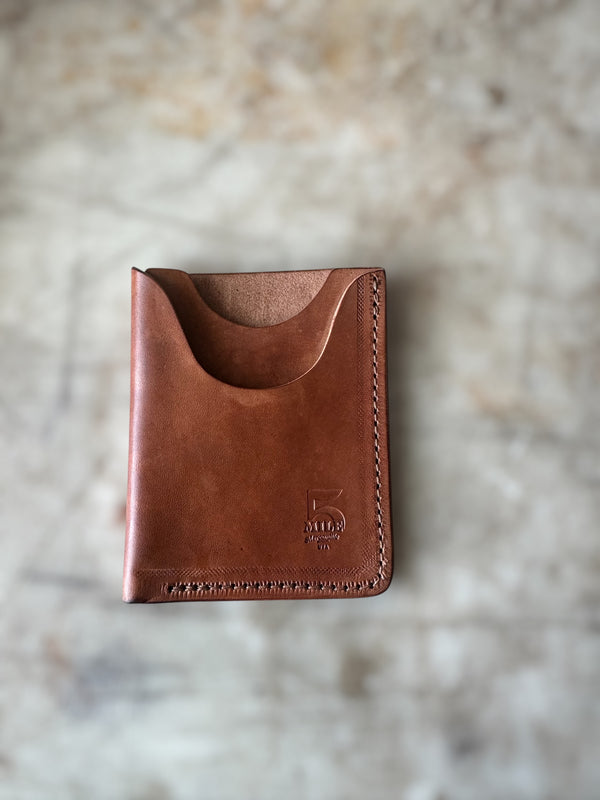 A brown leather card holder wallet with a curved top card slot, shown closed on a textured, light beige stone surface. The brand name 5 Mile is embossed near the bottom right corner.