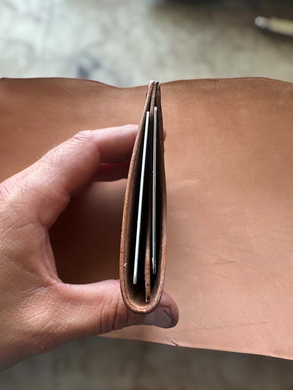 A close-up of a hand holding a slim brown leather wallet or card holder, showing several metallic cards inserted inside, against a tan leather background.