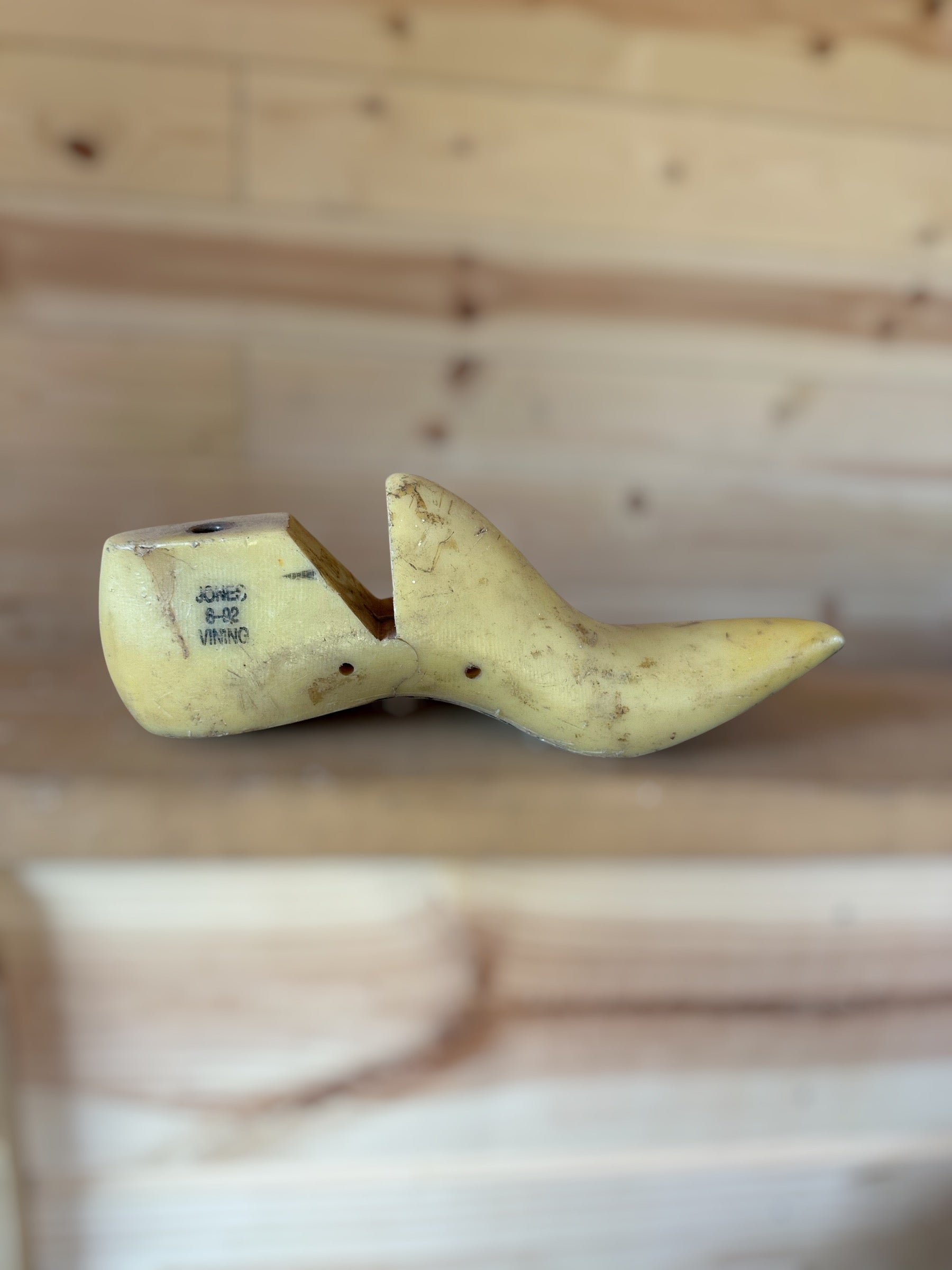 A single pale yellow wooden shoe tree with printed size markings, resting on a light wood surface with a matching blurred wood plank background.