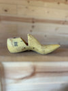 A single pale yellow wooden shoe tree with printed size markings, resting on a light wood surface with a matching blurred wood plank background.