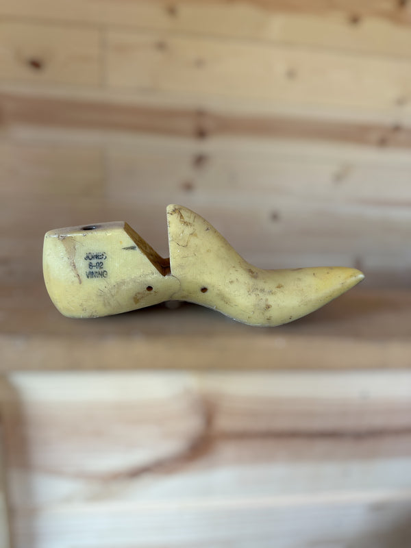 A single pale yellow wooden shoe tree with printed size markings, resting on a light wood surface with a matching blurred wood plank background.