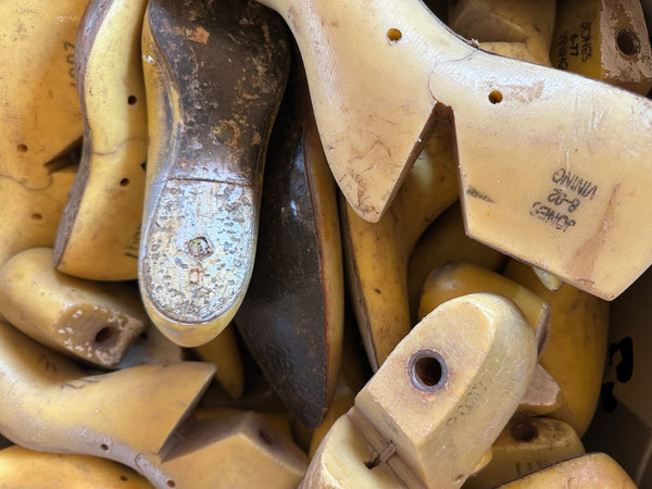 A close-up pile of vintage wooden shoe lasts, showing natural wood grain, some with stamped measurements and one with a dark sole.