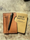 A tan leather card holder wallet with a pen in a side pocket and a Field Notes memo book tucked inside, photographed on a textured stone surface.