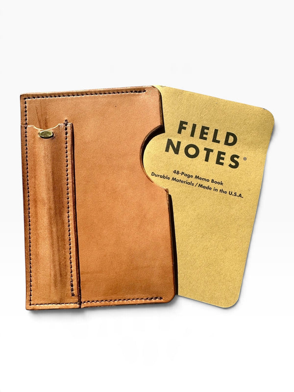 A tan leather Field Notes card holder wallet shown open, revealing a gold-toned 48-page memo book tucked inside. The leather has visible stitching and a small brass snap detail on the left side.