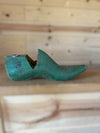 A green wooden shoe tree with a peg hole, resting on a light wood surface against a wooden plank background.