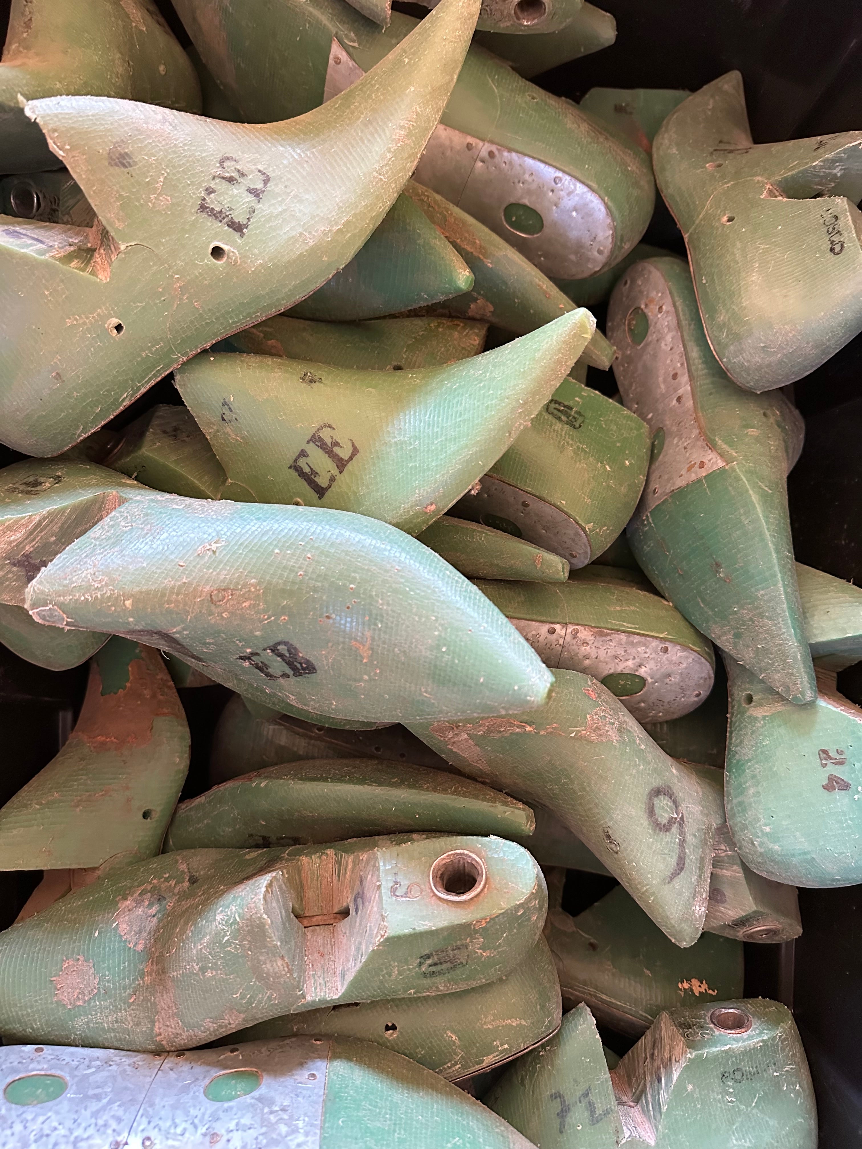 A pile of used, vintage shoe lasts made of molded wood, painted in a distressed sage green color with natural wood showing through, and some hand-written size markings.