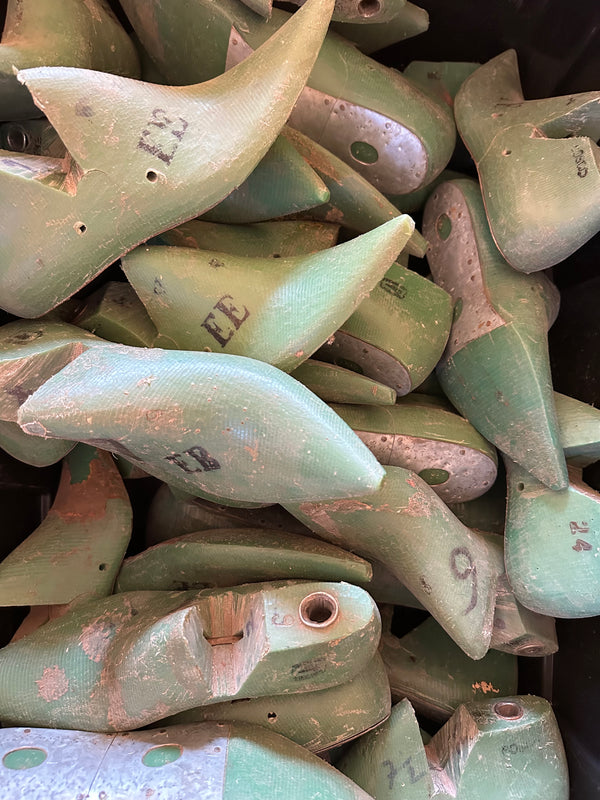 A pile of used, vintage shoe lasts made of molded wood, painted in a distressed sage green color with natural wood showing through, and some hand-written size markings.