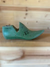 A vintage green wooden shoe tree with black printed numbers, resting on a light wood surface against a wooden background.