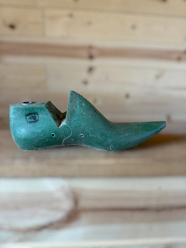 A vintage green wooden shoe tree with a pointed toe and a small black logo stamp, resting on a light wood plank surface.
