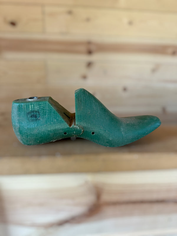 A vintage wooden shoe tree with a painted green finish, resting on a light wood surface against a matching wooden plank background.