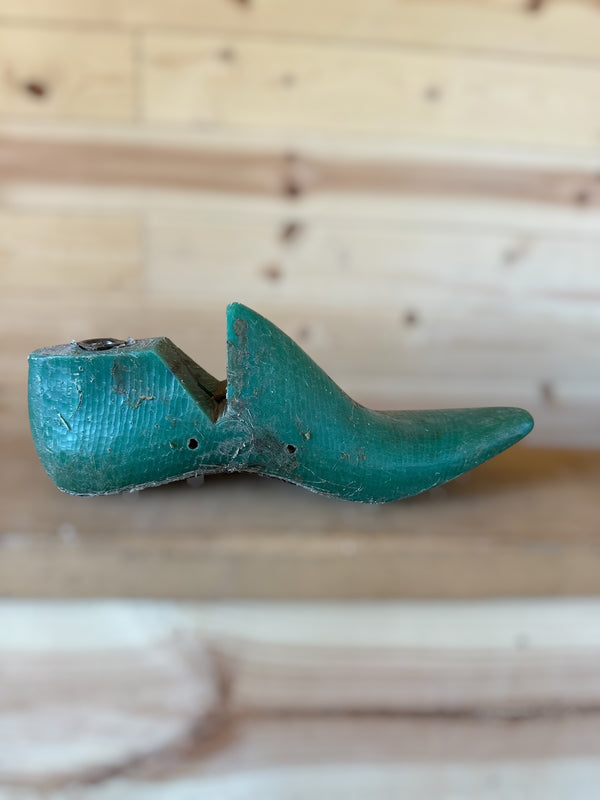 A vintage wooden shoe tree with a distressed teal-green painted finish, resting on a light wood plank surface against a matching wooden background.