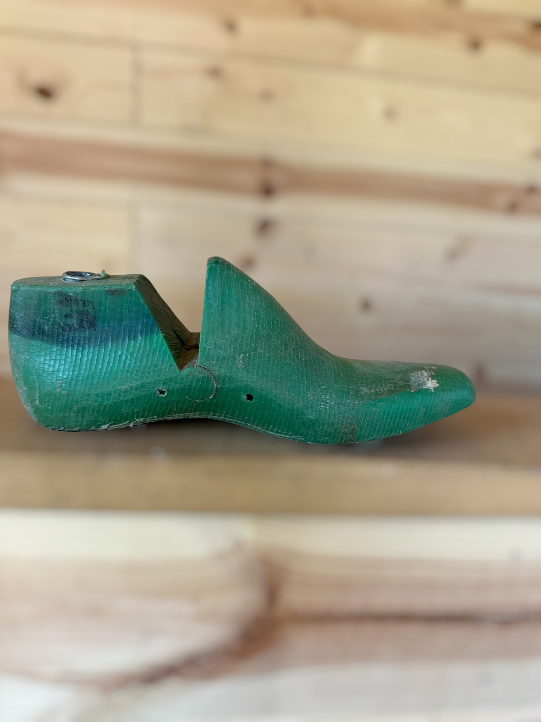A vintage green wooden shoe tree with a weathered, rustic finish, resting on a wooden surface against a light wood plank background.
