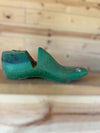A vintage green wooden shoe tree with a weathered, rustic finish, resting on a wooden surface against a light wood plank background.