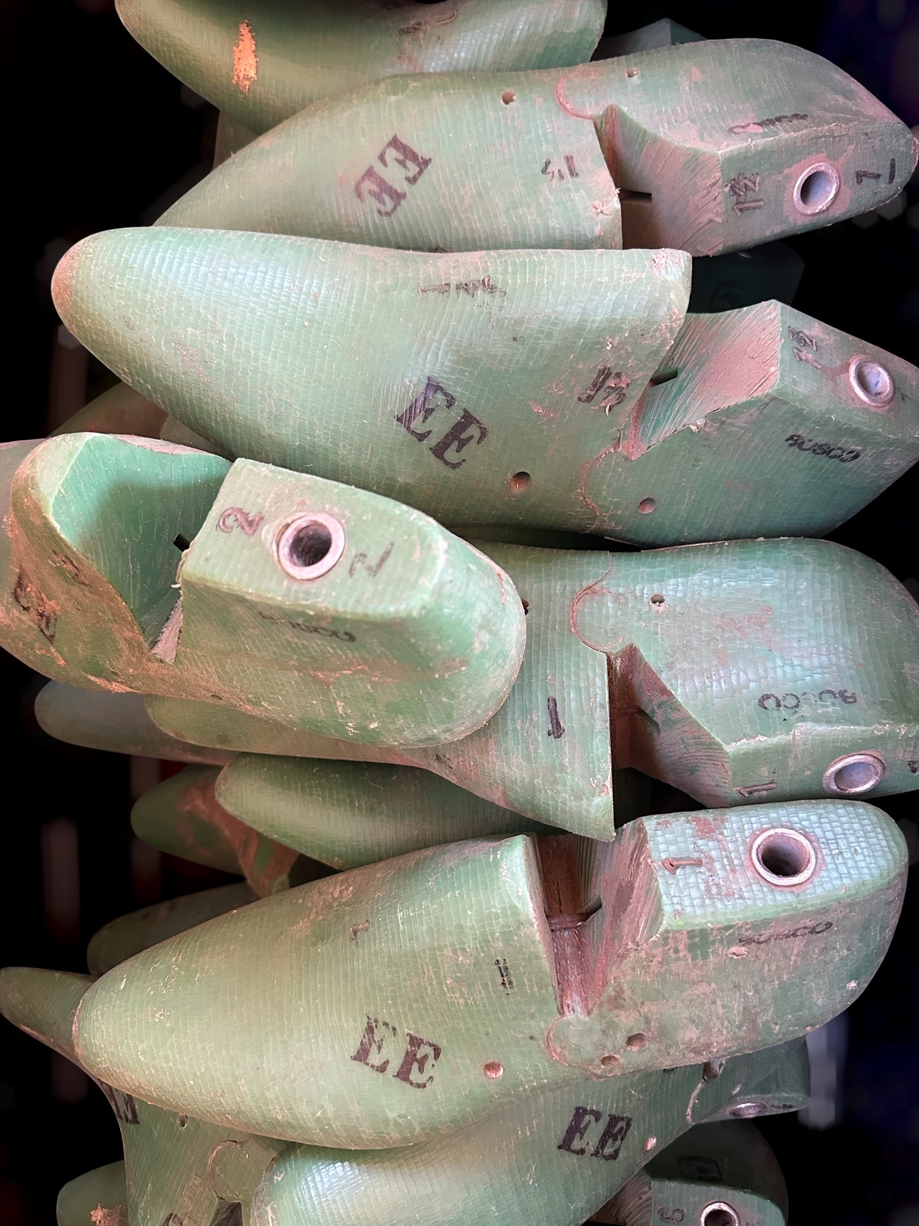 A close-up of a stack of mint green wooden shoe lasts with pinkish-red wear marks and stamped letters.