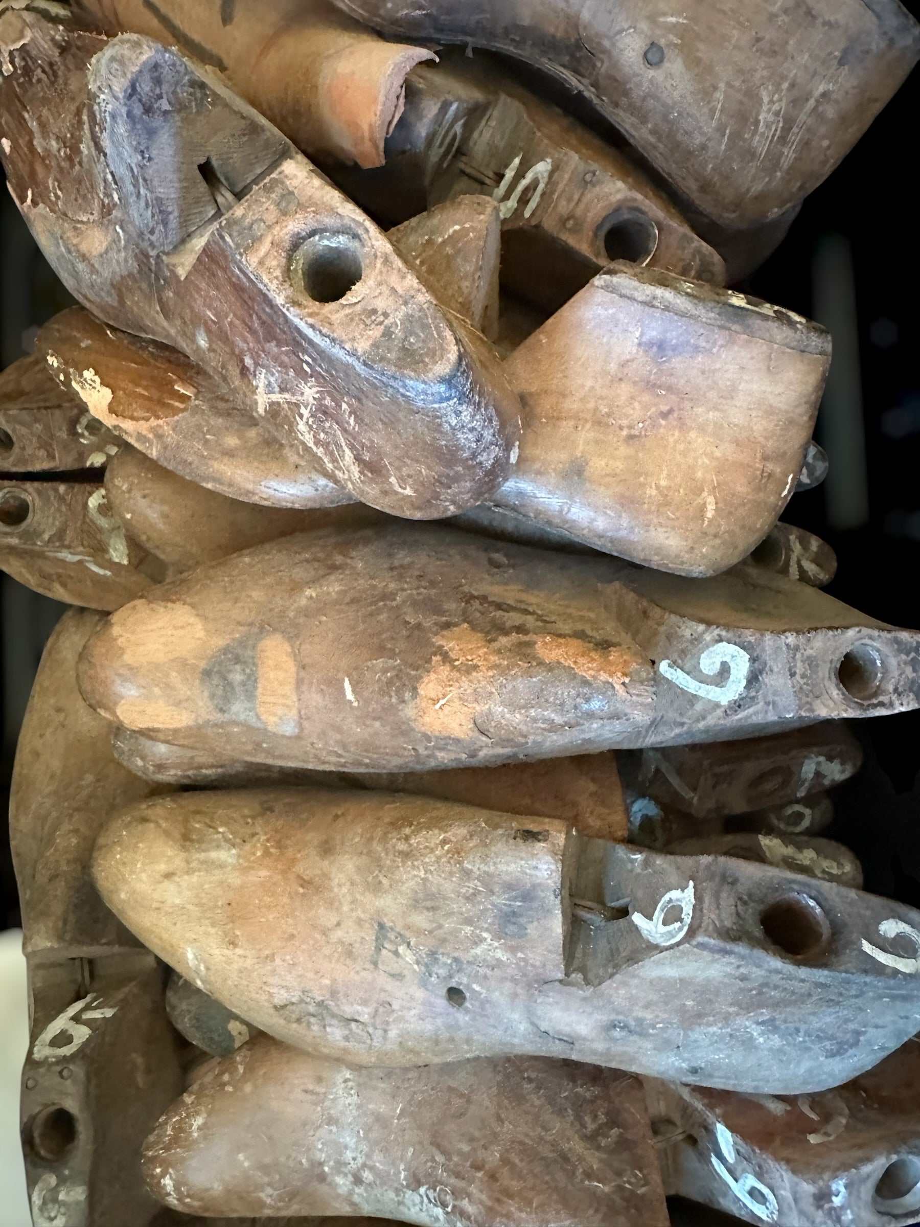 A close-up pile of vintage wooden shoe lasts, showing weathered brown wood with white handwritten numbers on some pieces.