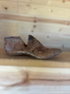 A single vintage wooden shoe tree with a weathered, rustic brown finish, resting on a light wooden surface against a matching wooden plank background.