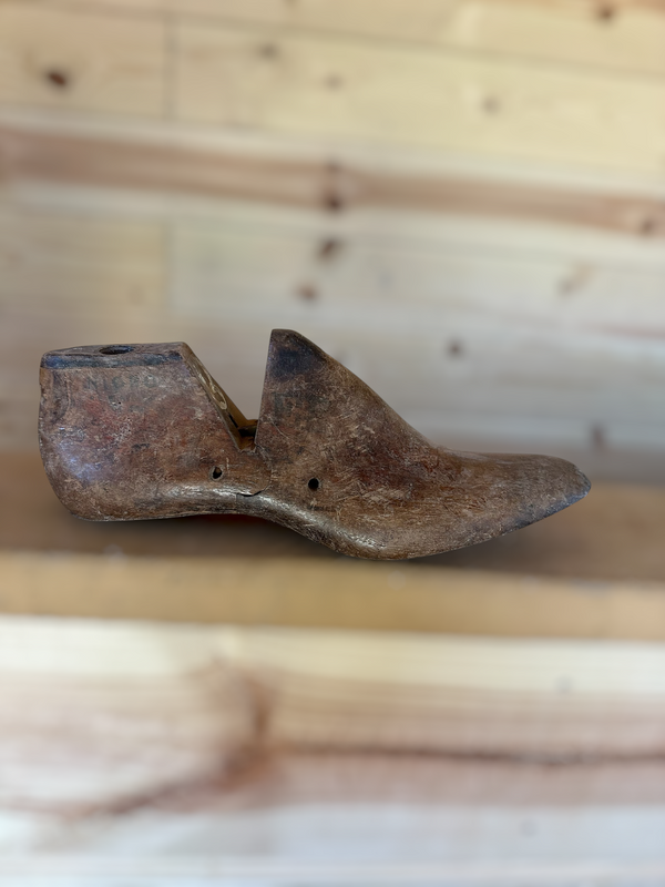 A single vintage wooden shoe tree with a weathered, rustic brown finish, resting on a light wooden surface against a matching wooden plank background.