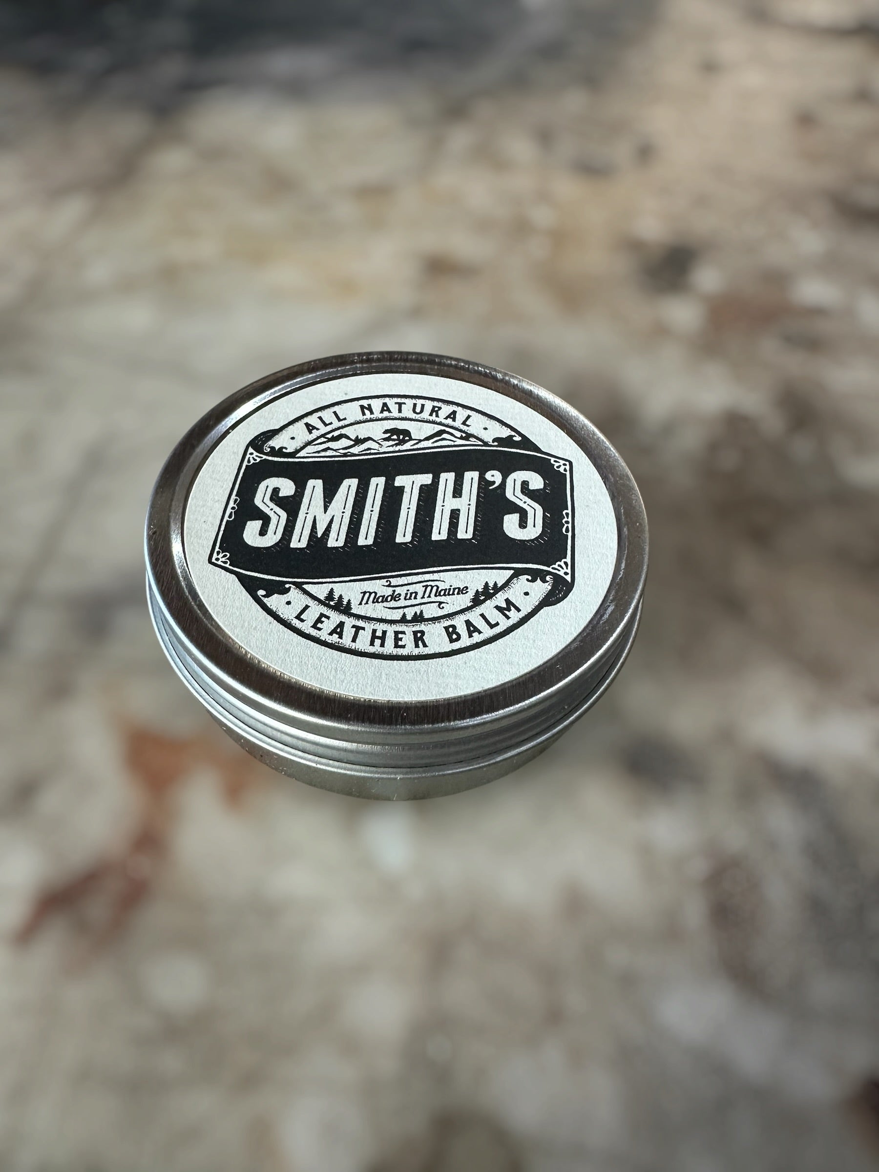 A round metal tin of Smith's Leather Balm with a white and black label, resting on a blurred stone surface.