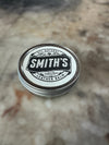 A round metal tin of Smith's Leather Balm with a white and black label, resting on a blurred stone surface.