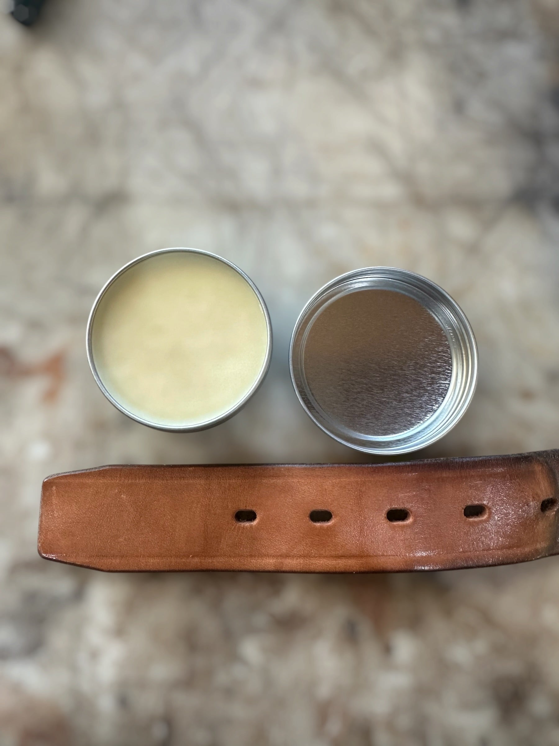 A small round metal tin of cream-colored balm or wax shown open next to a brown leather strap on a mottled stone-like surface.