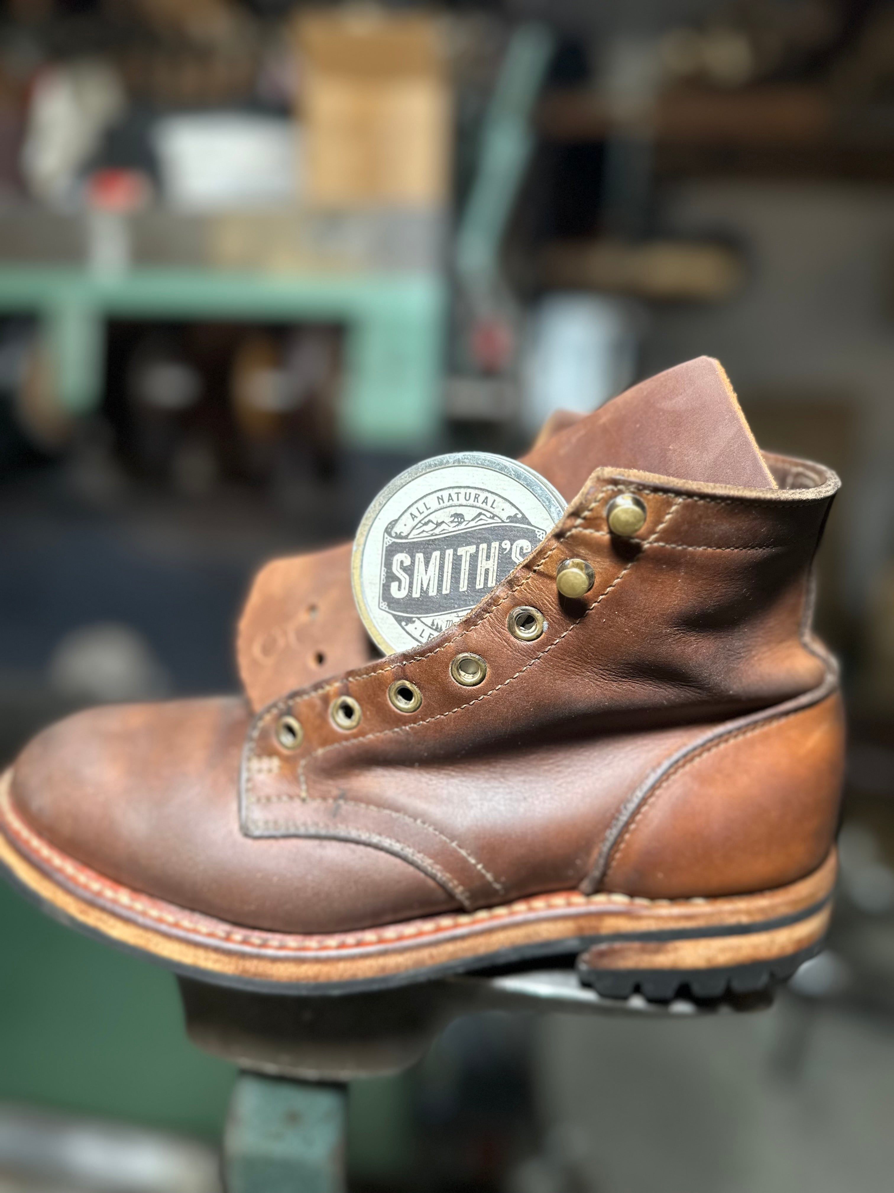 A brown leather work boot with a circular Smith's brand tag attached near the collar, photographed in a workshop setting with a blurred background.