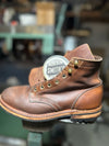 A brown leather work boot with a circular Smith's brand tag attached near the collar, photographed in a workshop setting with a blurred background.