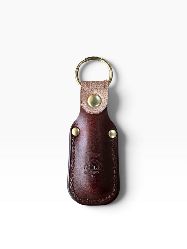 A dark brown leather key fob case with brass hardware, featuring a stamped logo on the front, shown on a clean white background.