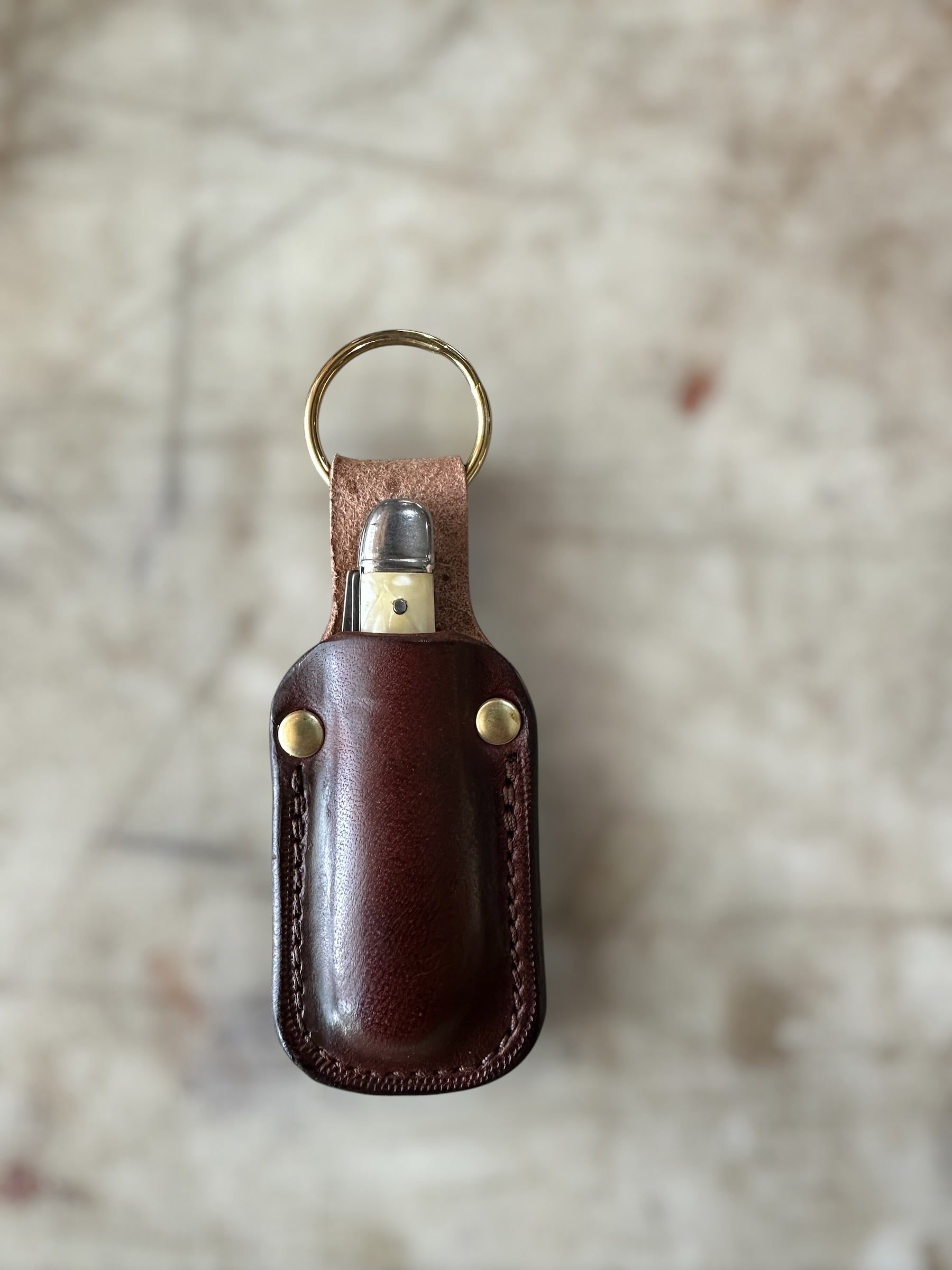 A dark brown leather lighter case with brass hardware, including a split key ring, two brass rivets, and a lighter peeking out from the top, set against a blurred light beige stone background.